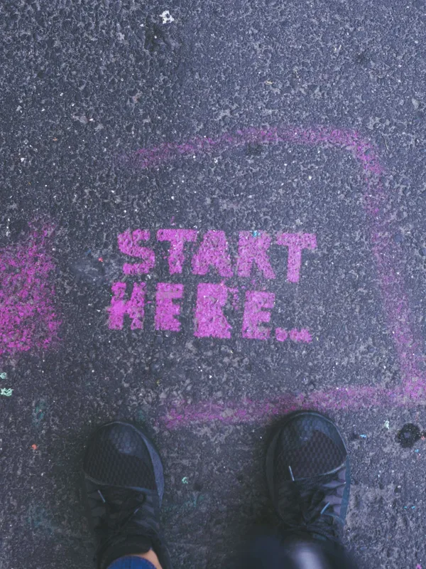 Pinke Schablonenschrift „Start here“ auf Asphalt, davor zwei Schuhspitzen